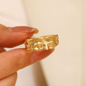 Bague visage Vénus dorée tenue entre deux doigts